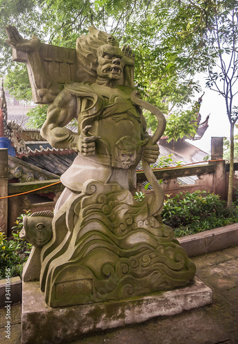 Fengdu, China - May 8, 2010: Ghost City, historic sanctuary. Closeup of brown stone bow holding muscular man-like angry monster statue on wall under green foliage and silver sky.