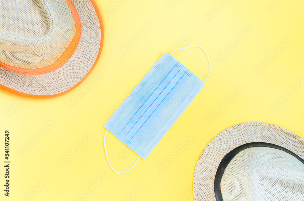 Obraz premium Stay home, social distancing, summer vacation travel concept due to global pandemic coronavirus. Two straw hats separated by medical mask on yellow background. Flat lay, top view, copy space.