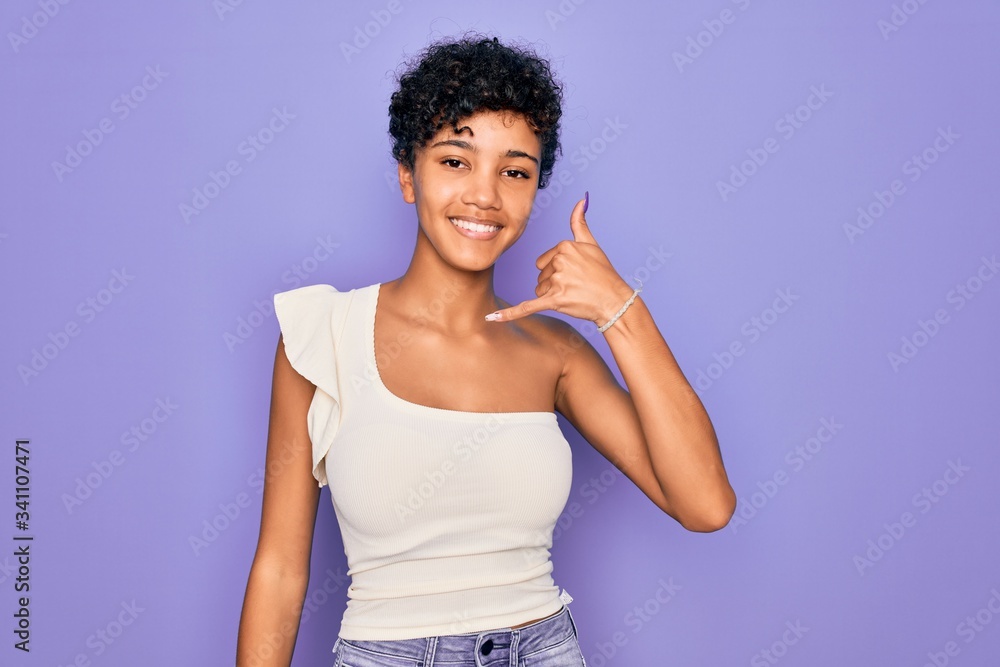 Young beautiful african american afro woman wearing casual t-shirt over purple background smiling doing phone gesture with hand and fingers like talking on the telephone. Communicating concepts. Young beautiful african american afro woman wearing casual t-shirt over purple background smiling doing phone gesture with hand and fingers like talking on the telephone. Communicating concepts.