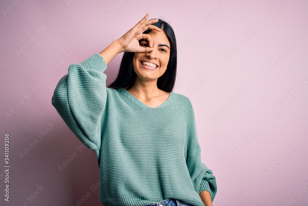 Young beautiful hispanic fashion woman wearing green sweater over pink background doing ok gesture with hand smiling, eye looking through fingers with happy face. Young beautiful hispanic fashion woman wearing green sweater over pink background doing ok gesture with hand smiling, eye looking through fingers with happy face.