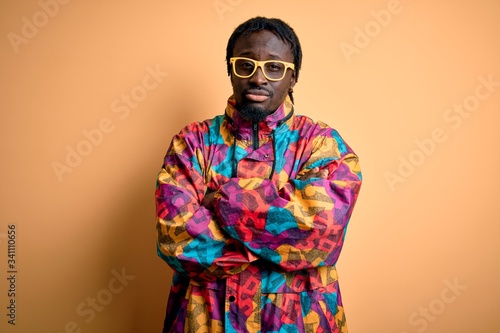 Handsome african american man wearing colorful coat and glasses over yellow background skeptic and nervous, disapproving expression on face with crossed arms. Negative person.