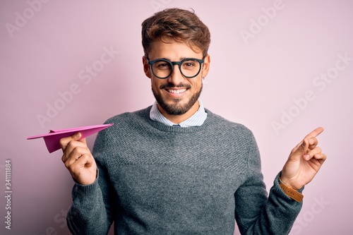 Young handsome man holding paper aircraft wearing glasses over isolated pink background very happy pointing with hand and finger to the side