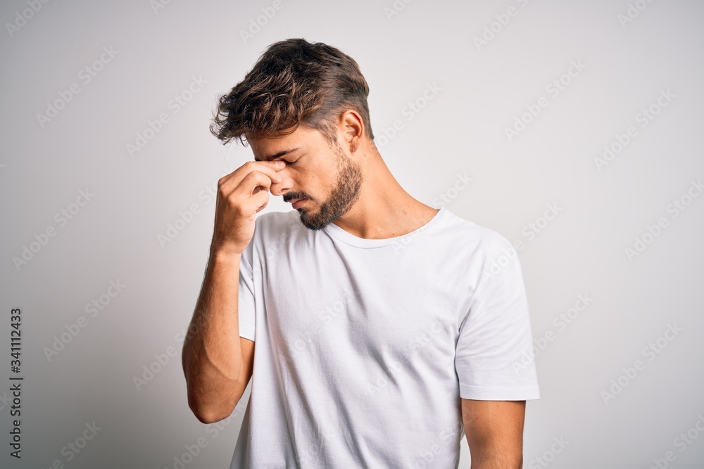 Young handsome man with beard wearing casual t-shirt standing over white background tired rubbing nose and eyes feeling fatigue and headache. Stress and frustration concept. Young handsome man with beard wearing casual t-shirt standing over white background tired rubbing nose and eyes feeling fatigue and headache. Stress and frustration concept.