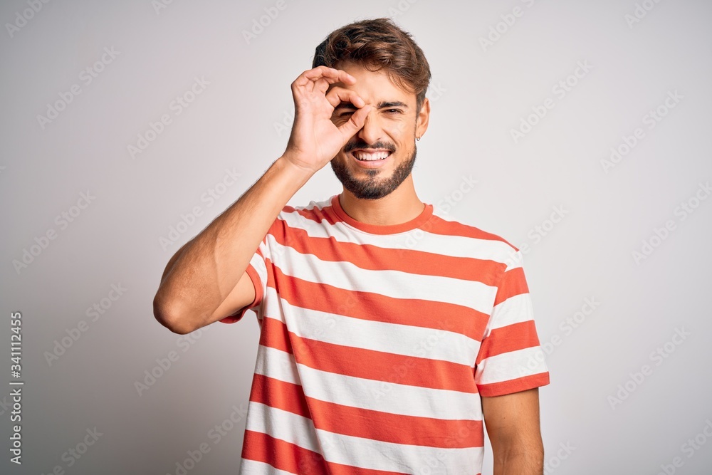 Young handsome man with beard wearing striped t-shirt standing over white background doing ok gesture with hand smiling, eye looking through fingers with happy face. Young handsome man with beard wearing striped t-shirt standing over white background doing ok gesture with hand smiling, eye looking through fingers with happy face.