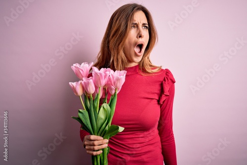 Young beautiful brunette woman holding bouquet of pink tulips over isolated background angry and mad screaming frustrated and furious, shouting with anger. Rage and aggressive concept.