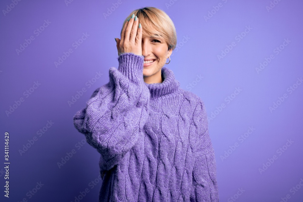 Young blonde woman with short hair wearing winter turtleneck sweater over purple background covering one eye with hand, confident smile on face and surprise emotion. Young blonde woman with short hair wearing winter turtleneck sweater over purple background covering one eye with hand, confident smile on face and surprise emotion.