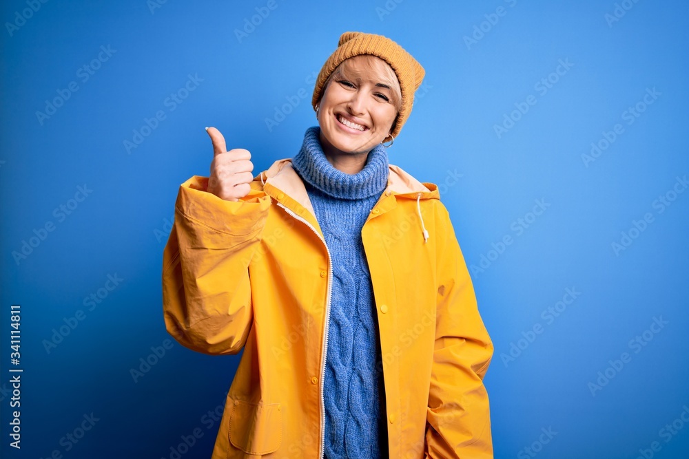 Young blonde woman with short hair wearing rain coat for rainy weather over blue background doing happy thumbs up gesture with hand. Approving expression looking at the camera showing success. Young blonde woman with short hair wearing rain coat for rainy weather over blue background doing happy thumbs up gesture with hand. Approving expression looking at the camera showing success.