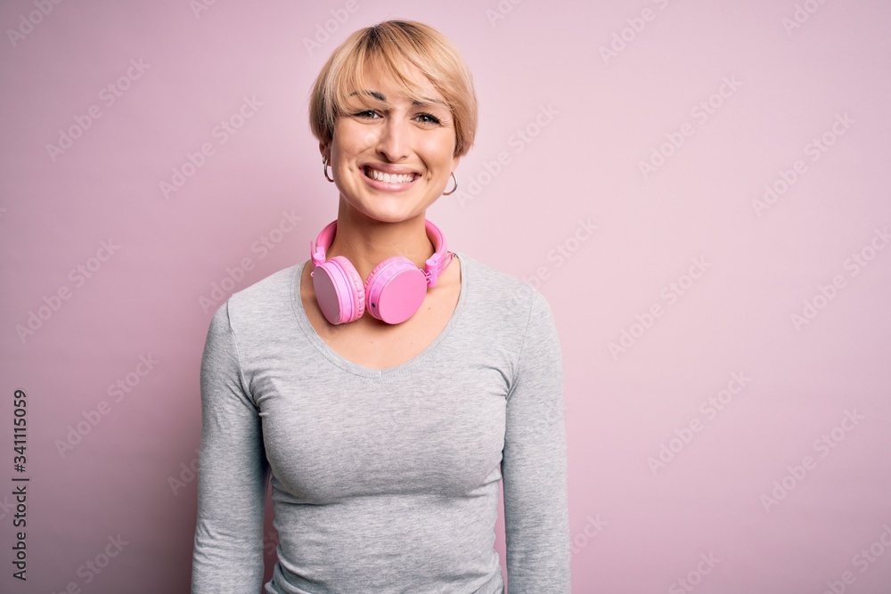 Young blonde woman with short hair wearing headphones on neck over pink background with a happy and cool smile on face. Lucky person. Young blonde woman with short hair wearing headphones on neck over pink background with a happy and cool smile on face. Lucky person.