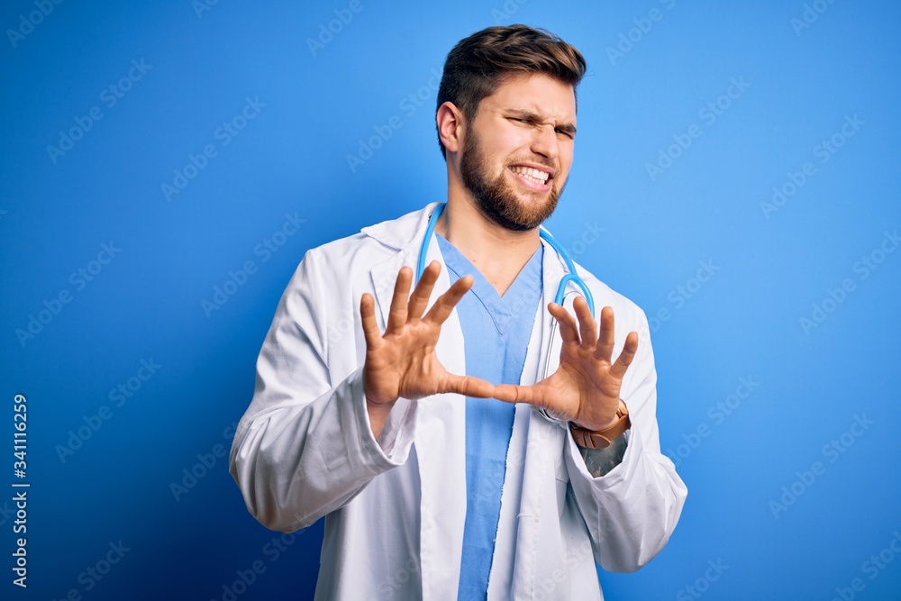 Young blond doctor man with beard and blue eyes wearing white coat and stethoscope disgusted expression, displeased and fearful doing disgust face because aversion reaction. With hands raised Young blond doctor man with beard and blue eyes wearing white coat and stethoscope disgusted expression, displeased and fearful doing disgust face because aversion reaction. With hands raised