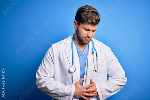 Young blond doctor man with beard and blue eyes wearing white coat and stethoscope with hand on stomach because indigestion, painful illness feeling unwell. Ache concept.