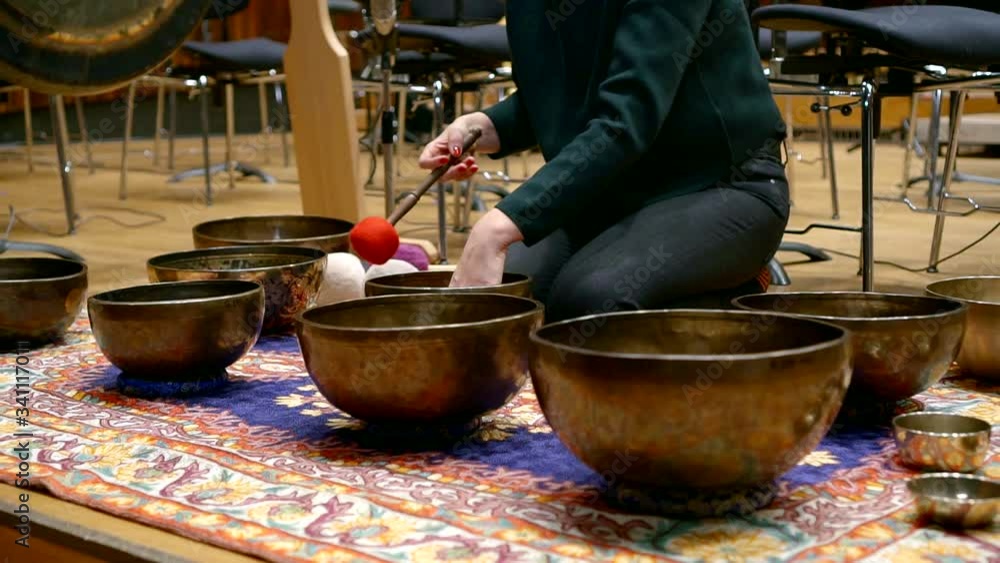a woman in black clothes is sitting on her knees on the carpet. she strikes the singing bowls with her stick. the camera moves away a woman in black clothes is sitting on her knees on the carpet. she strikes the singing bowls with her stick. the camera moves away