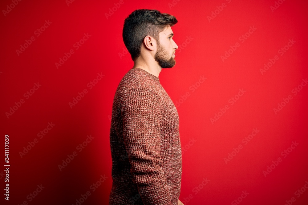 Young blond man with beard and blue eyes wearing casual sweater over red background looking to side, relax profile pose with natural face with confident smile. Young blond man with beard and blue eyes wearing casual sweater over red background looking to side, relax profile pose with natural face with confident smile.