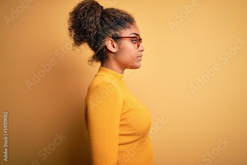 Young beautiful african american girl wearing sweater and glasses over yellow background looking to side, relax profile pose with natural face and confident smile.