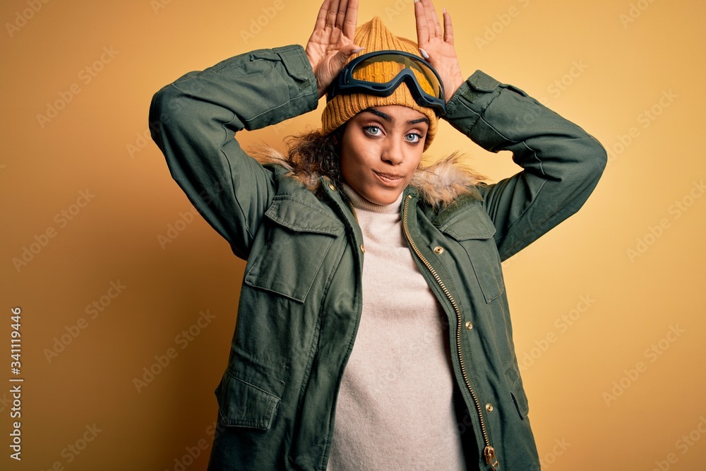Young african american afro skier girl wearing snow sportswear and ski goggles Doing bunny ears gesture with hands palms looking cynical and skeptical. Easter rabbit concept. Young african american afro skier girl wearing snow sportswear and ski goggles Doing bunny ears gesture with hands palms looking cynical and skeptical. Easter rabbit concept.