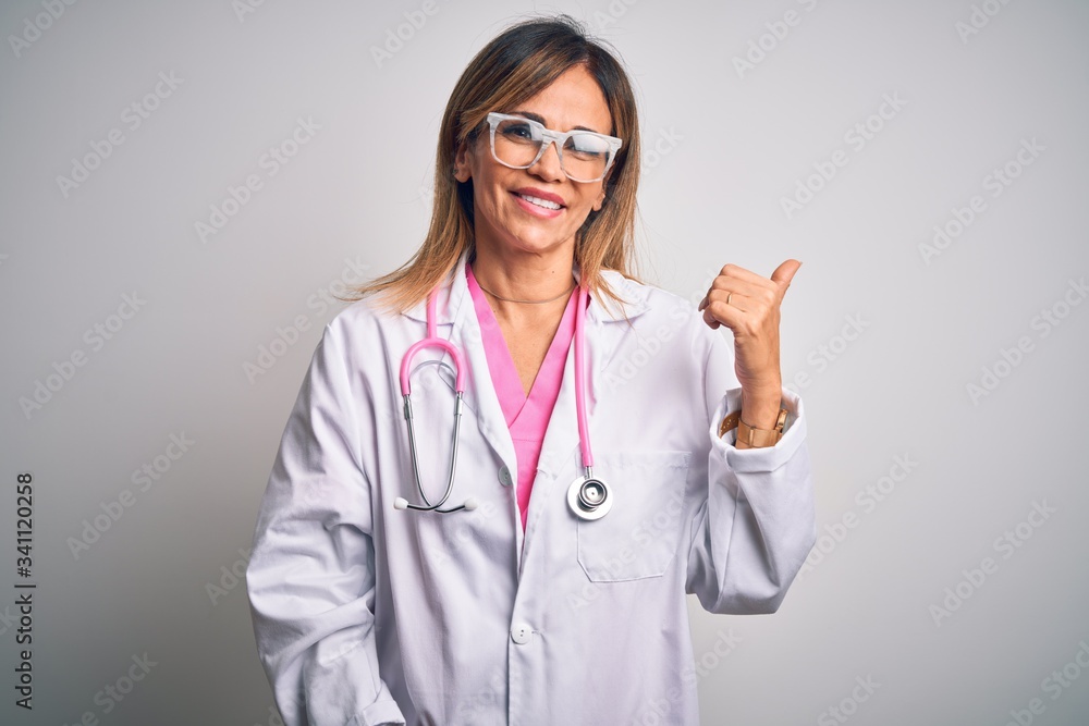 Middle age beautiful doctor woman wearing pink stethoscope over isolated white background smiling with happy face looking and pointing to the side with thumb up.