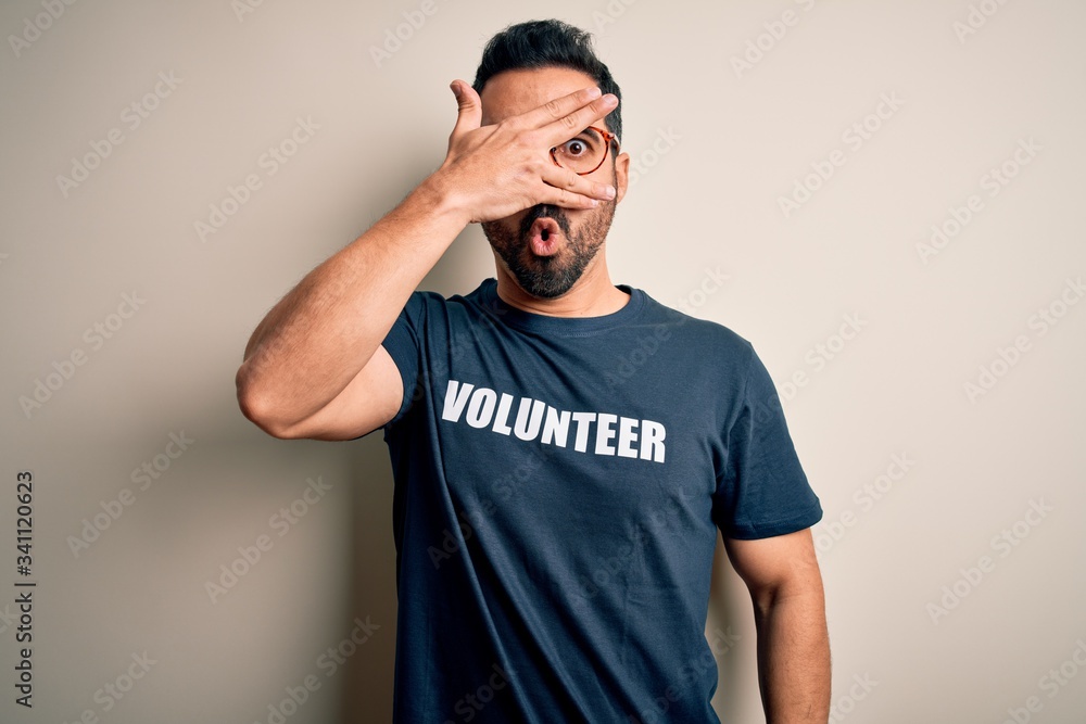 Handsome man with beard wearing t-shirt with volunteer message over white background peeking in shock covering face and eyes with hand, looking through fingers with embarrassed expression. Handsome man with beard wearing t-shirt with volunteer message over white background peeking in shock covering face and eyes with hand, looking through fingers with embarrassed expression.