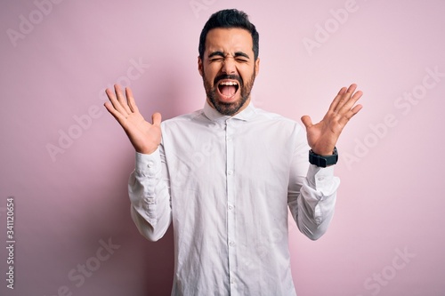Young handsome man with beard wearing casual shirt standing over pink background celebrating mad and crazy for success with arms raised and closed eyes screaming excited. Winner concept © Krakenimages.com