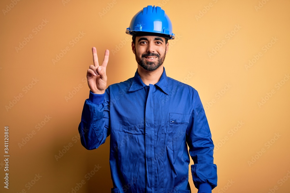 Mechanic man with beard wearing blue uniform and safety helmet over yellow background smiling looking to the camera showing fingers doing victory sign. Number two.