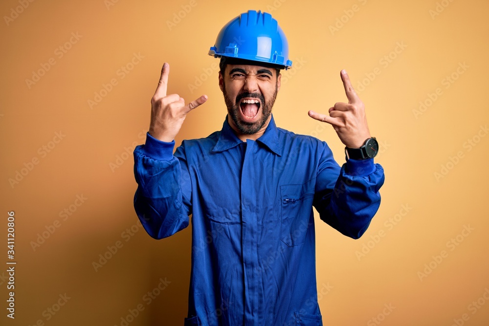 Mechanic man with beard wearing blue uniform and safety helmet over yellow background shouting with crazy expression doing rock symbol with hands up. Music star. Heavy music concept.
