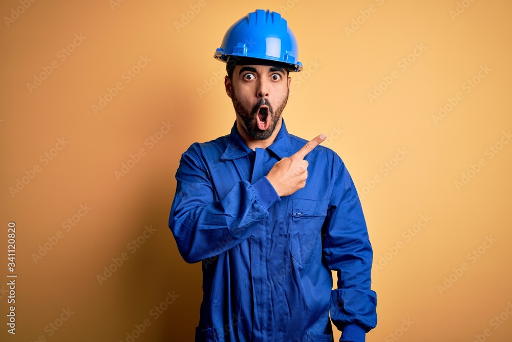 Mechanic man with beard wearing blue uniform and safety helmet over yellow background Surprised pointing with finger to the side, open mouth amazed expression.