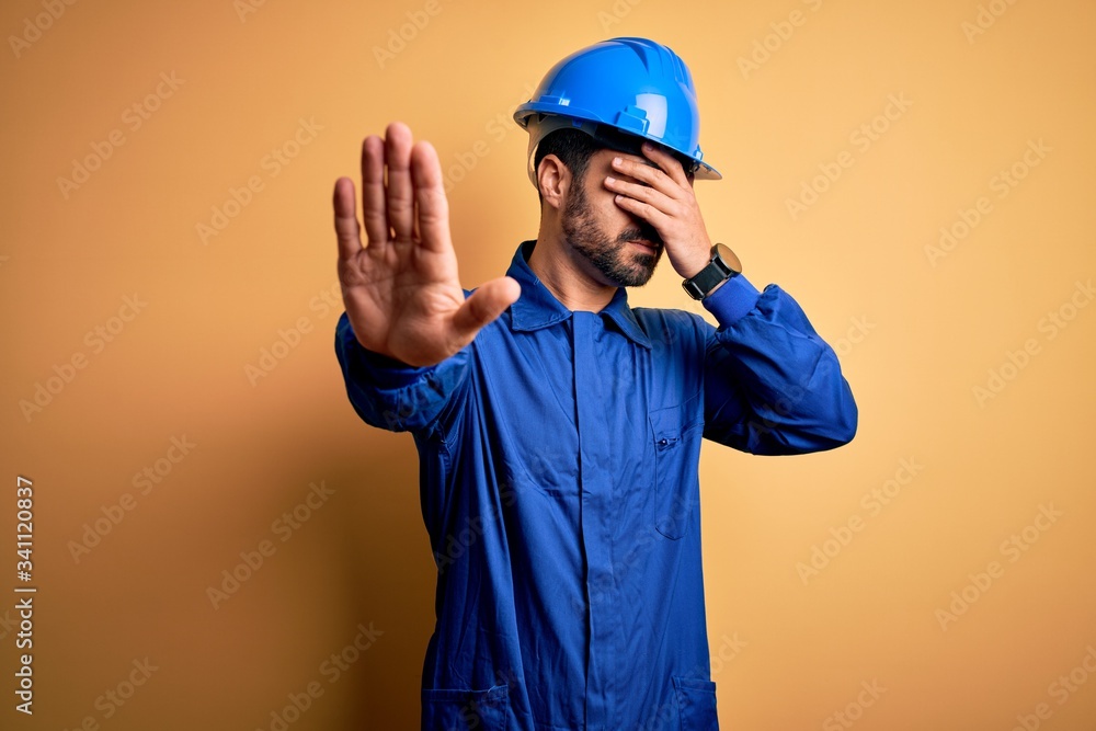 Mechanic man with beard wearing blue uniform and safety helmet over yellow background covering eyes with hands and doing stop gesture with sad and fear expression. Embarrassed and negative concept.