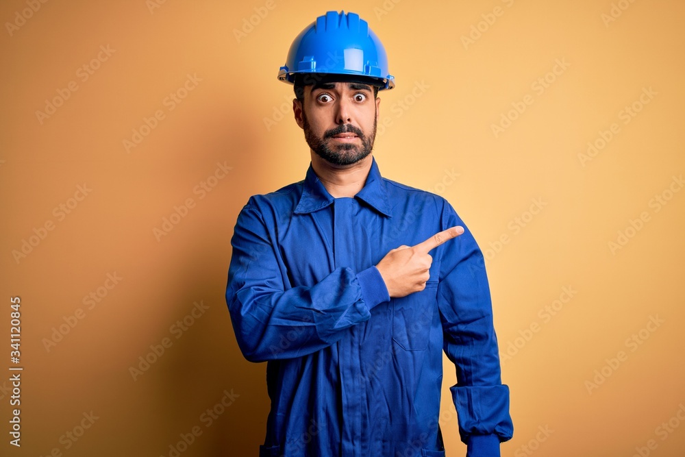 Mechanic man with beard wearing blue uniform and safety helmet over yellow background Pointing aside worried and nervous with forefinger, concerned and surprised expression