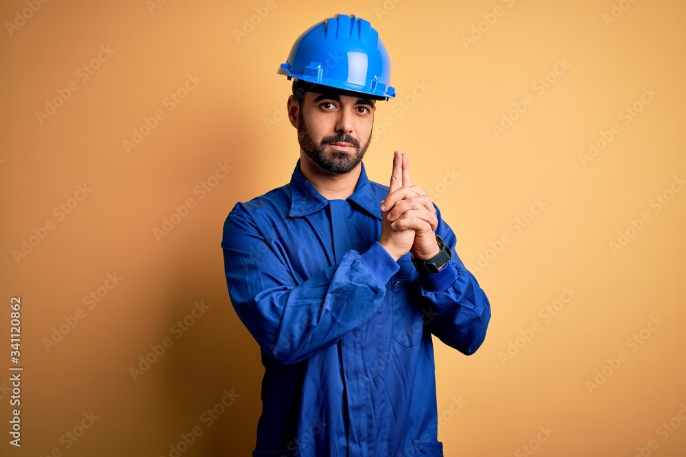 Mechanic man with beard wearing blue uniform and safety helmet over yellow background Holding symbolic gun with hand gesture, playing killing shooting weapons, angry face