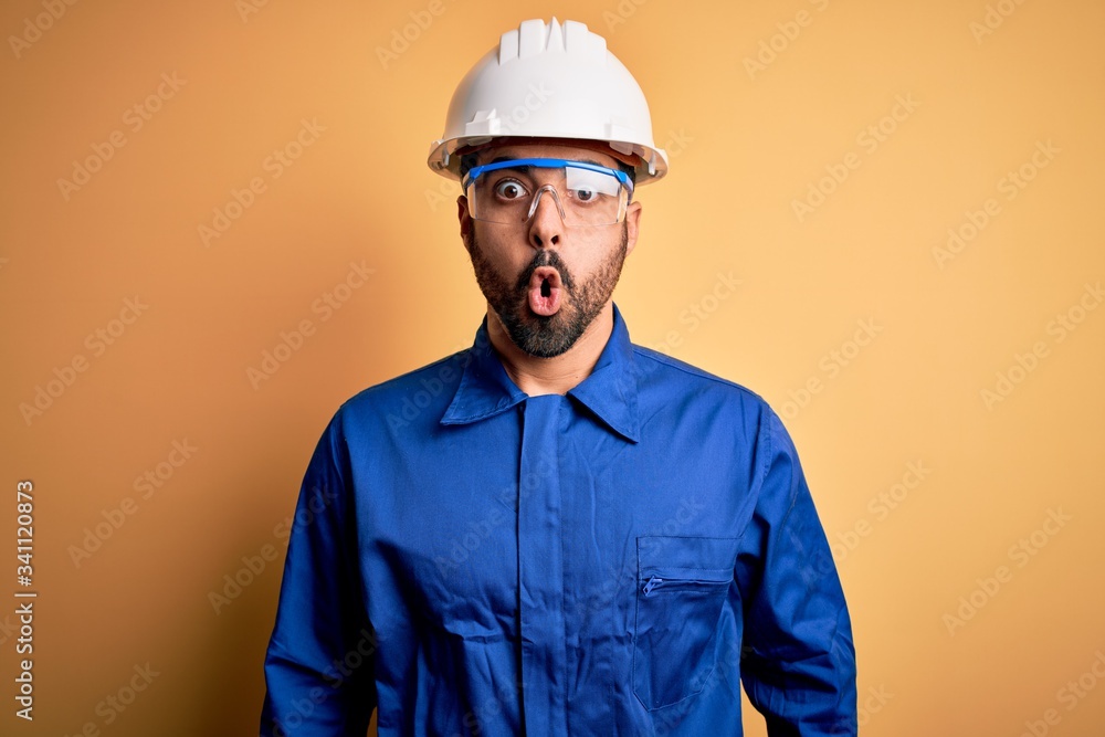 Mechanic man with beard wearing blue uniform and safety glasses over yellow background afraid and shocked with surprise expression, fear and excited face.