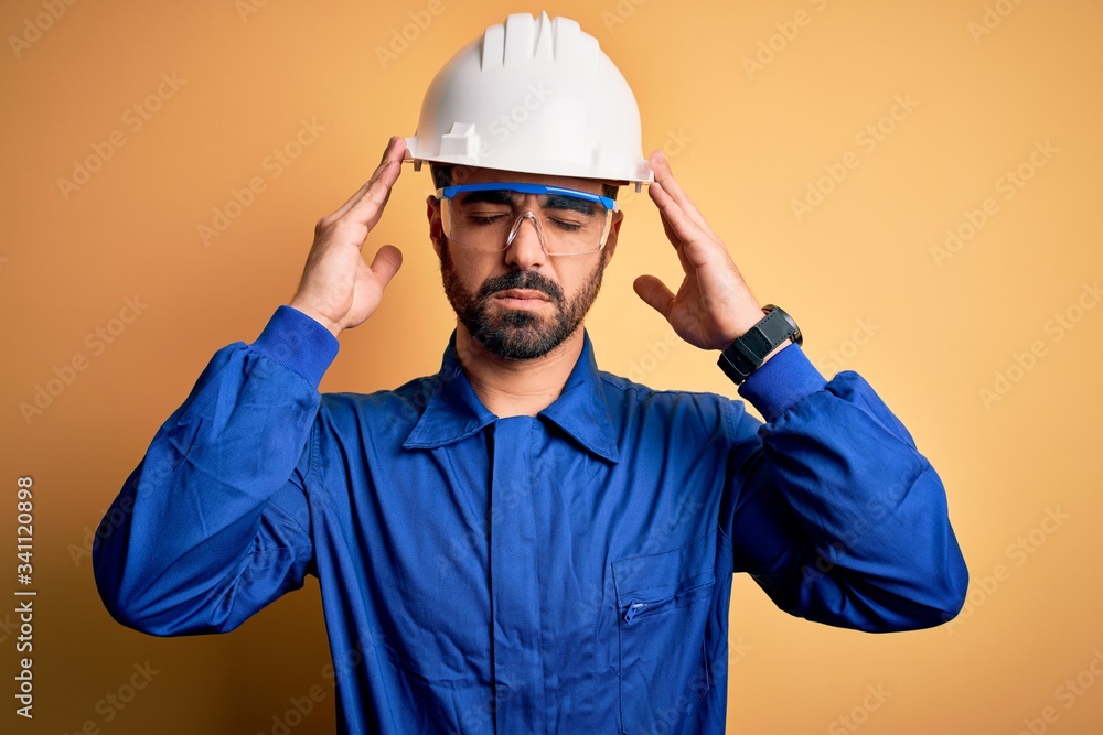 Mechanic man with beard wearing blue uniform and safety glasses over yellow background suffering from headache desperate and stressed because pain and migraine. Hands on head.