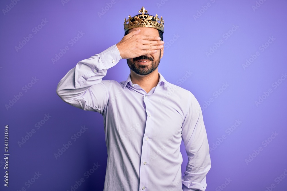 Young handsome man with beard wearing golden crown of king over purple background smiling and laughing with hand on face covering eyes for surprise. Blind concept. Young handsome man with beard wearing golden crown of king over purple background smiling and laughing with hand on face covering eyes for surprise. Blind concept.