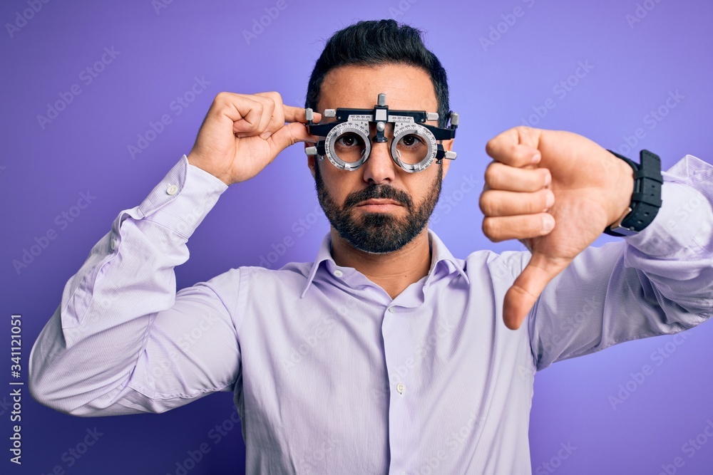 Young handsome optical man with beard wearing optometry glasses over purple background with angry face, negative sign showing dislike with thumbs down, rejection concept Young handsome optical man with beard wearing optometry glasses over purple background with angry face, negative sign showing dislike with thumbs down, rejection concept