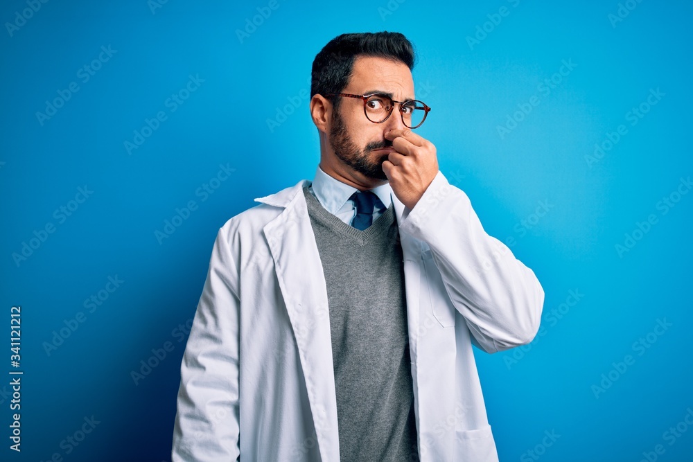 Young handsome doctor man with beard wearing coat and glasses over blue background smelling something stinky and disgusting, intolerable smell, holding breath with fingers on nose. Bad smell Young handsome doctor man with beard wearing coat and glasses over blue background smelling something stinky and disgusting, intolerable smell, holding breath with fingers on nose. Bad smell