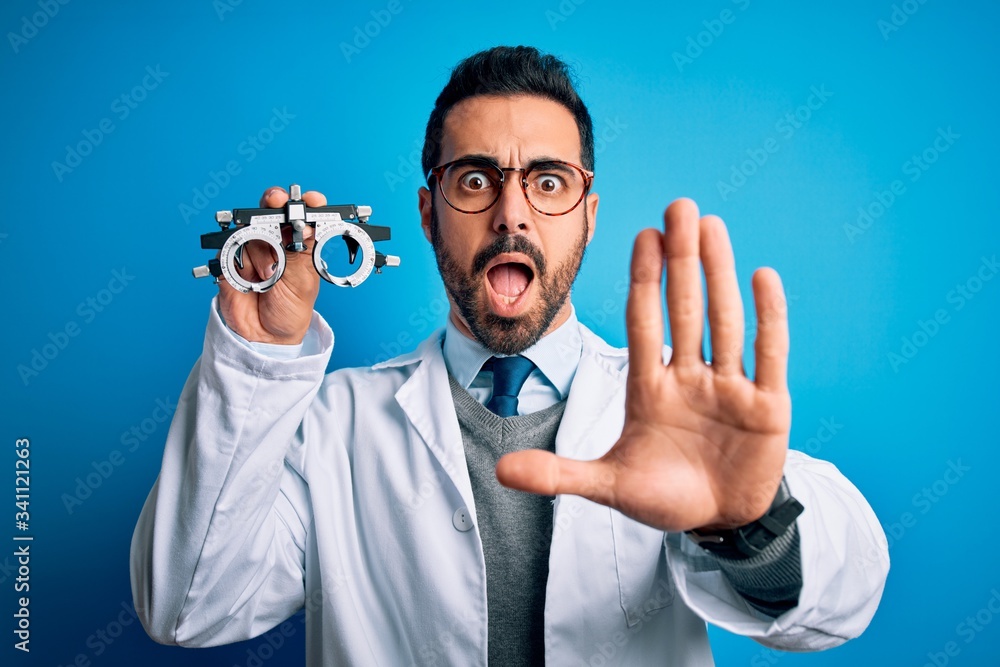 Naklejka premium Young handsome optical man with beard holding optometry glasses over blue background doing stop gesture with hands palms, angry and frustration expression