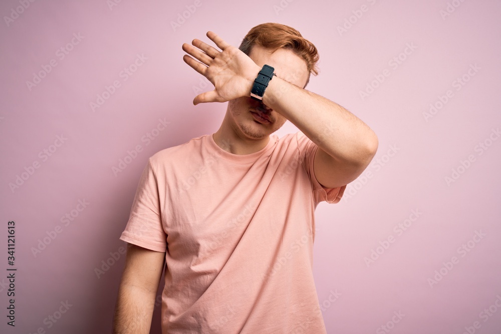 Young handsome redhead man wearing casual t-shirt standing over isolated pink background covering eyes with arm, looking serious and sad. Sightless, hiding and rejection concept Young handsome redhead man wearing casual t-shirt standing over isolated pink background covering eyes with arm, looking serious and sad. Sightless, hiding and rejection concept