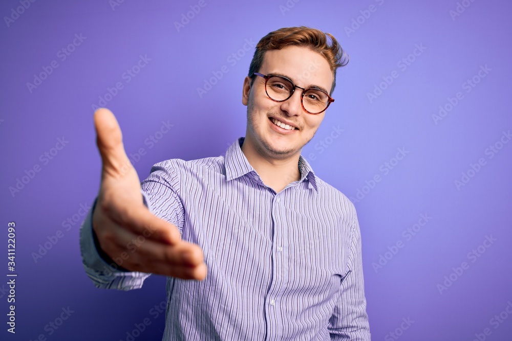 Naklejka premium Young handsome redhead man wearing casual shirt and glasses over purple background smiling friendly offering handshake as greeting and welcoming. Successful business.