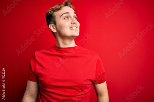 Young handsome redhead man wearing casual t-shirt over isolated red background looking away to side with smile on face, natural expression. Laughing confident. © Krakenimages.com