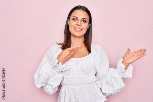 Young beautiful brunette woman wearing casual white dress standing over pink background amazed and smiling to the camera while presenting with hand and pointing with finger. © Krakenimages.com
