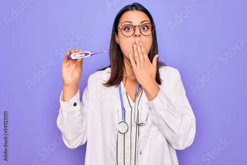Young beautiful brunette doctor woman wearing stethoscope and coat holding thermometer covering mouth with hand, shocked and afraid for mistake. Surprised expression
