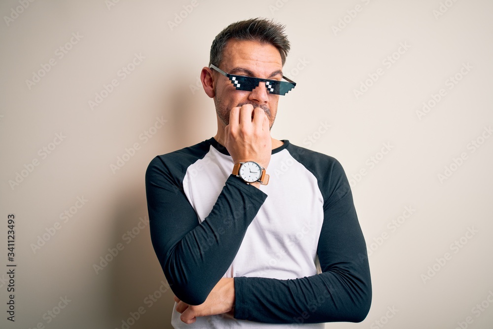 Young handsome man wearing funny thug life sunglasses meme over white background looking stressed and nervous with hands on mouth biting nails. Anxiety problem. Young handsome man wearing funny thug life sunglasses meme over white background looking stressed and nervous with hands on mouth biting nails. Anxiety problem.