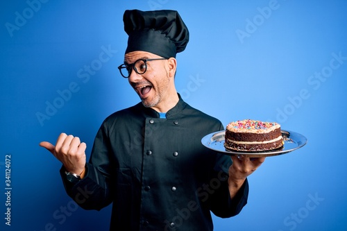 Young handsome baker man wearing cooker uniform and hat holding tray with cake pointing and showing with thumb up to the side with happy face smiling