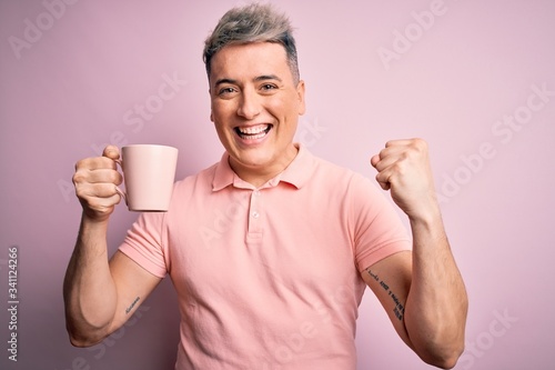 Young modern handsome man drinking a cup of hot coffee over isolated pink background screaming proud and celebrating victory and success very excited, cheering emotion © Krakenimages.com