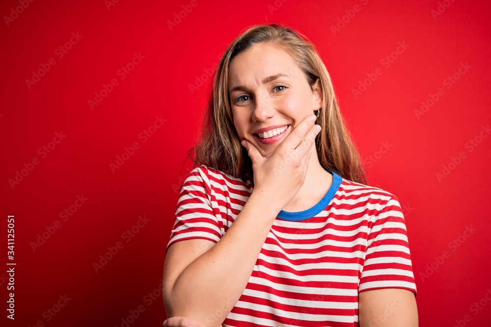 Young beautiful blonde woman wearing casual striped t-shirt over isolated red background looking confident at the camera smiling with crossed arms and hand raised on chin. Thinking positive.