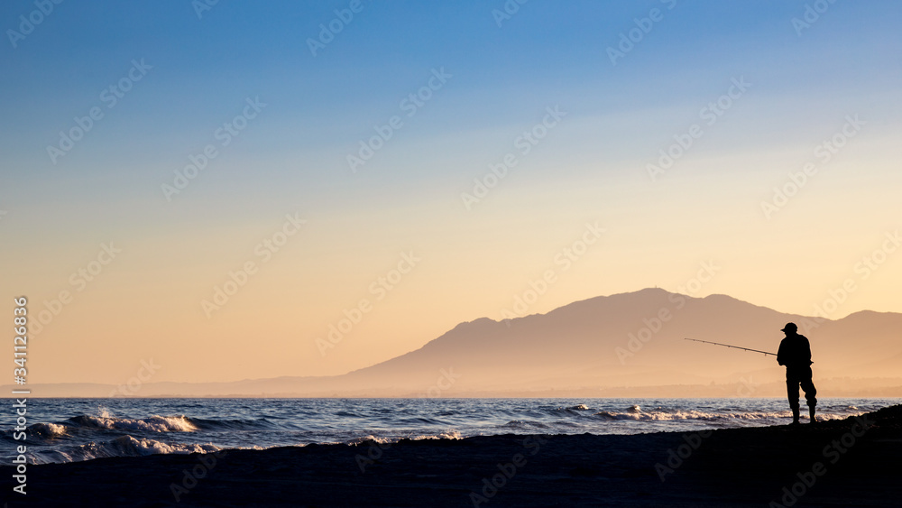 Backlit fisherman on Cabopino beach, Marbella, Malaga, with mountains in the background.