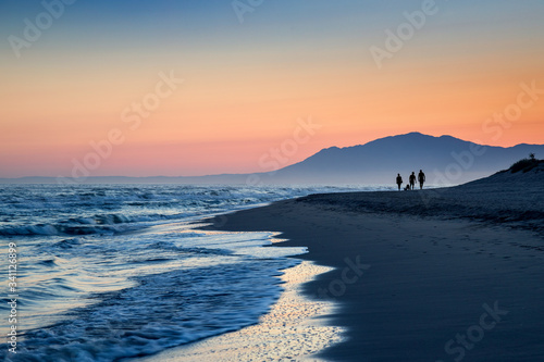 Three silhouettes of people stroll along the beach of Cabopino, Marbella, Malaga during sunset with mountains in the background.