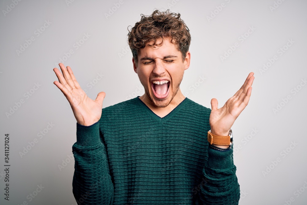 Young blond handsome man with curly hair wearing green sweater over white background celebrating mad and crazy for success with arms raised and closed eyes screaming excited. Winner concept Young blond handsome man with curly hair wearing green sweater over white background celebrating mad and crazy for success with arms raised and closed eyes screaming excited. Winner concept