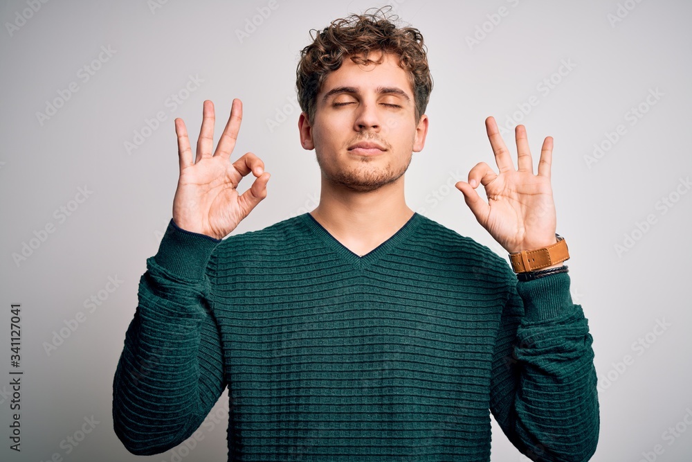 Young blond handsome man with curly hair wearing green sweater over white background relax and smiling with eyes closed doing meditation gesture with fingers. Yoga concept. Young blond handsome man with curly hair wearing green sweater over white background relax and smiling with eyes closed doing meditation gesture with fingers. Yoga concept.
