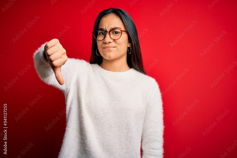 Young beautiful asian woman wearing casual sweater and glasses over red background looking unhappy and angry showing rejection and negative with thumbs down gesture. Bad expression.