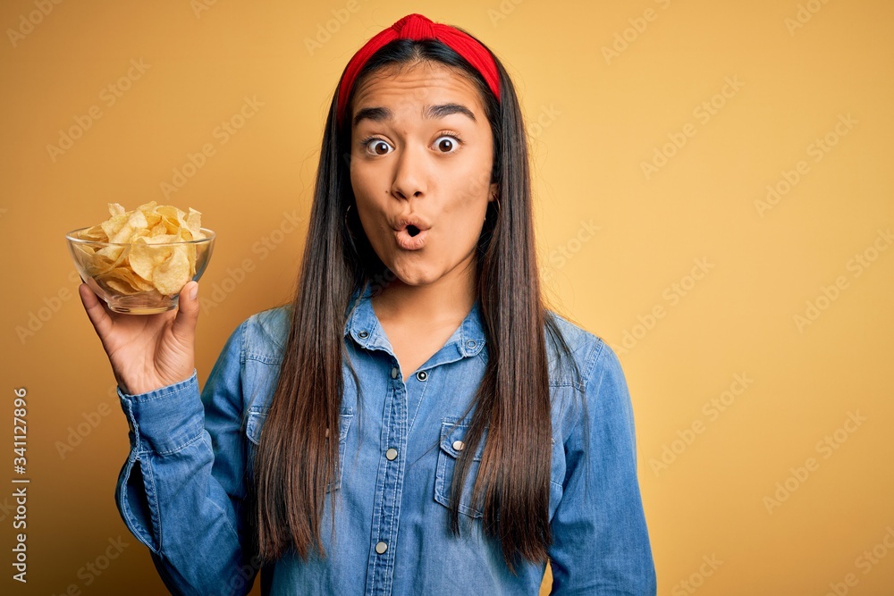 Young beautiful asian woman holding bowl with chips potatoes over isolated yellow background scared in shock with a surprise face, afraid and excited with fear expression Young beautiful asian woman holding bowl with chips potatoes over isolated yellow background scared in shock with a surprise face, afraid and excited with fear expression
