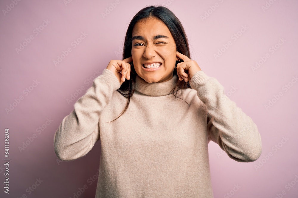Young beautiful asian woman wearing casual turtleneck sweater over pink background covering ears with fingers with annoyed expression for the noise of loud music. Deaf concept. Young beautiful asian woman wearing casual turtleneck sweater over pink background covering ears with fingers with annoyed expression for the noise of loud music. Deaf concept.