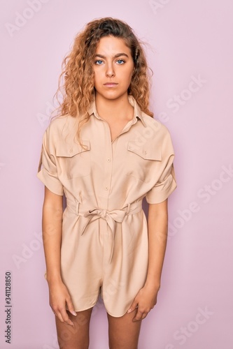 Young beautiful woman with blue eyes wearing casual summer dress over pink background with serious expression on face. Simple and natural looking at the camera.
